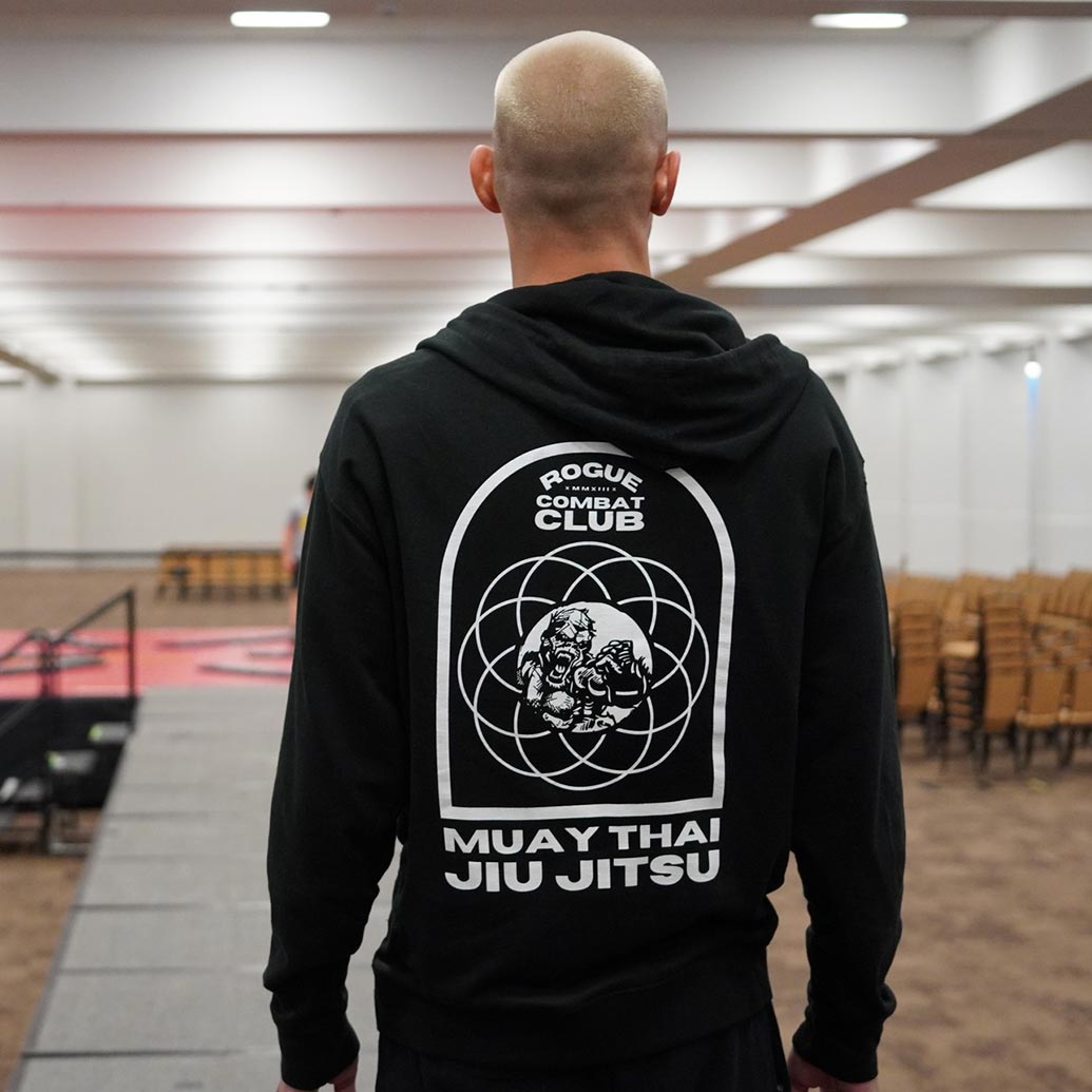 the back of a man with a bald hair cut and black hoodie with a logo of an aggressive gorilla in a circle with city skyline and the words Rogue Combat Club Muay Thai Jiu Jitsu in a conference room