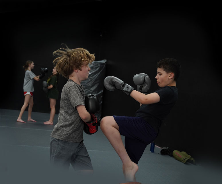 Young children practicing muay thai. One male child kicks another child who is blacking in the foreground and in the background there are two girls